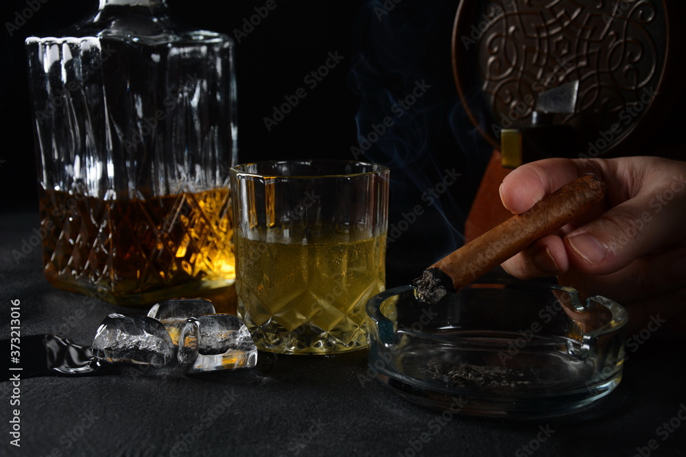 Glass of the whiskey with ice cubes and a square decanter, and cigar on a black background. Glass of the whiskey with ice cubes and a square decanter, and cigar on a black background.