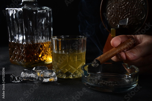 Glass of the whiskey with ice cubes and a square decanter, and cigar on a black background.