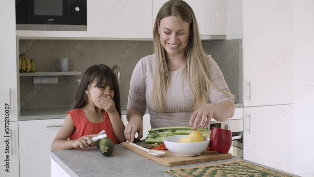 Cheerful mom cutting vegetables in kitchen, giving slice to daughter to taste, having fun with girl while cooking dinner together. Static shot. Family cooking concept Cheerful mom cutting vegetables in kitchen, giving slice to daughter to taste, having fun with girl while cooking dinner together. Static shot. Family cooking concept
