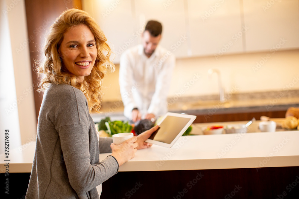 Woman using tablet at the kitchen table while man preparing food Woman using tablet at the kitchen table while man preparing food