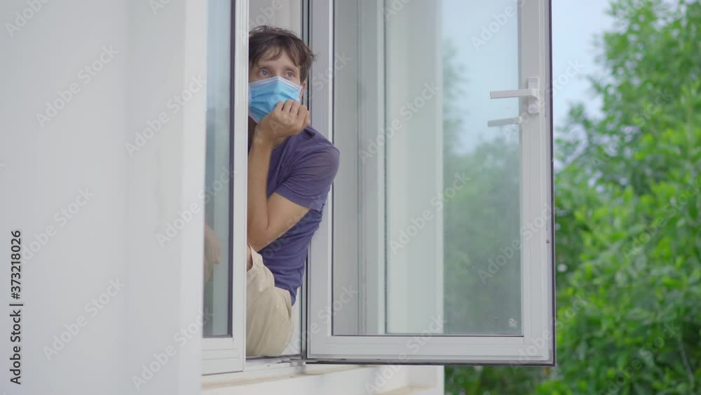 A young man in mandatory quarantine. He stays in one room for two weeks. He did not shave for a long time, he is stressed and upset. He sits by the window wearing a facemask and looks outside. Staying A young man in mandatory quarantine. He stays in one room for two weeks. He did not shave for a long time, he is stressed and upset. He sits by the window wearing a facemask and looks outside. Staying