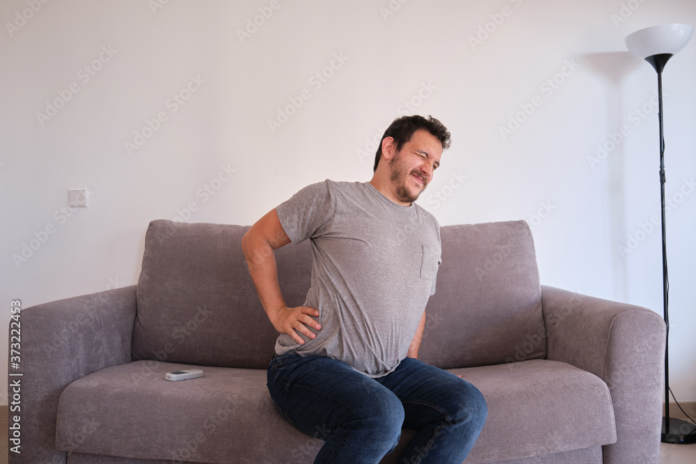 Young man sitting on a sofa suffering from low back pain. Backache concept. Young man sitting on a sofa suffering from low back pain. Backache concept.