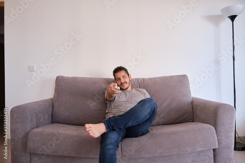 Portrait of a relaxed young man sitting on a sofa watching television and changing channels with the remote control on his hands. © Ladanifer