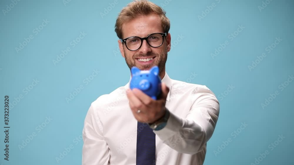 businessman holding a piggy bank in one hand while putting money with the other one and pointing at the camera on blue background businessman holding a piggy bank in one hand while putting money with the other one and pointing at the camera on blue background
