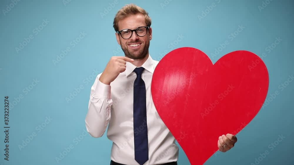 young sexy businessman holding a big red heart, pointing at the camera, at him and at the heart on blue background young sexy businessman holding a big red heart, pointing at the camera, at him and at the heart on blue background