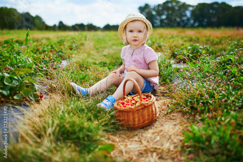 Adorable toddler girl in straw hat picking fresh organic strawberries on farm