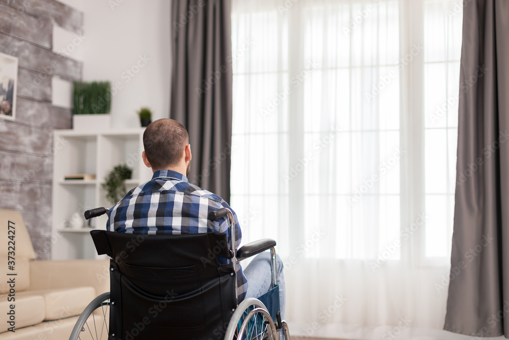 Unhappy man in wheelchair in living room looking at window. Serious sad caucasian man wearing casual clothes and look at large panoramic view in bright modern living room, hospital or clinic.