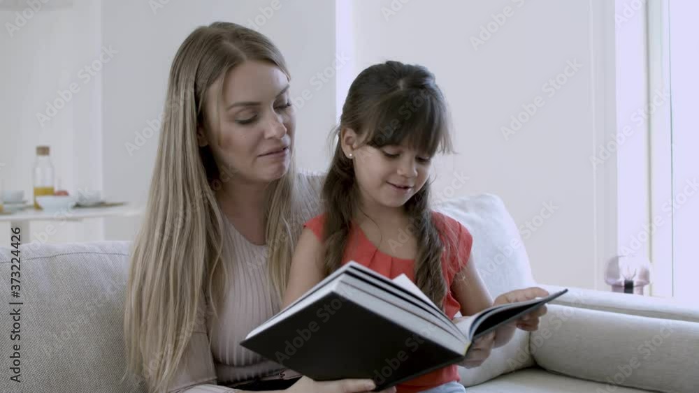 Little girl and her mom reading book together in living room. Cute daughter sitting on her mother lap. Medium shot. Childhood or literature concept Little girl and her mom reading book together in living room. Cute daughter sitting on her mother lap. Medium shot. Childhood or literature concept