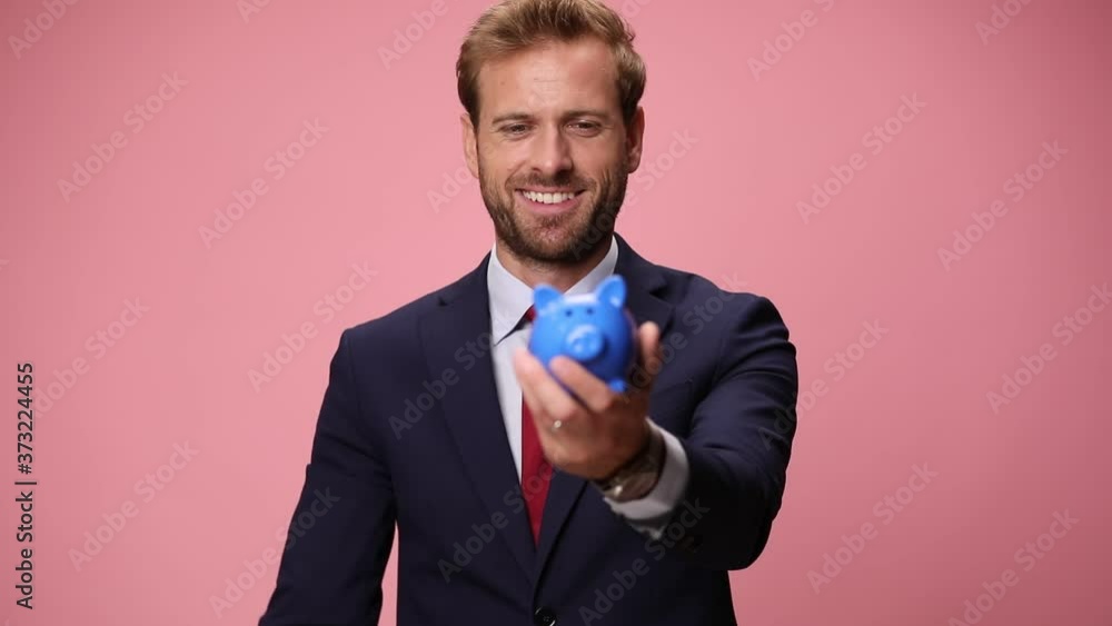 happy young guy holding piggy bank up, saving money by putting a coin and making thumbs up sign, pointing fingers on pink background happy young guy holding piggy bank up, saving money by putting a coin and making thumbs up sign, pointing fingers on pink background