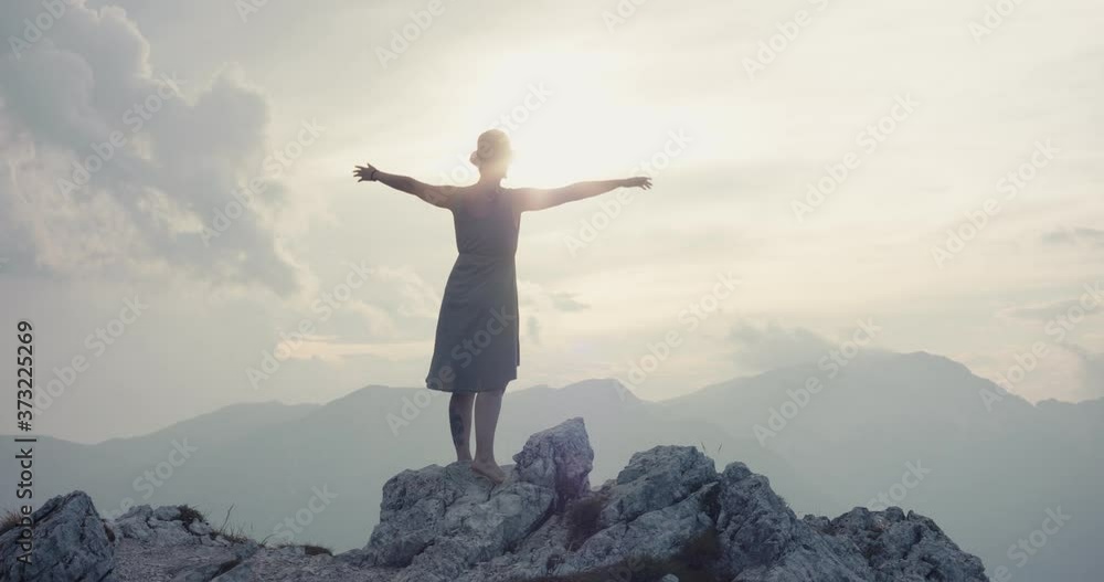 Woman wear summer dress hat on a hike in the mountains hiking in fall or autumn cloudy evening lift extend arms at the peak top rock at the view slowmotion light flare through cloud silhouette Woman wear summer dress hat on a hike in the mountains hiking in fall or autumn cloudy evening lift extend arms at the peak top rock at the view slowmotion light flare through cloud silhouette