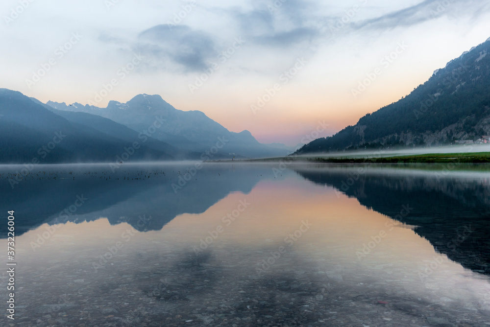 A fisherman rowing among the fog on the lake of Silvaplana in the Engadin valley at sunrise with mountains reflecting in the water