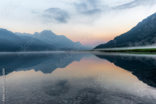 Fototapeta Naklejka Na Ścianę i Meble -  A fisherman rowing among the fog on the lake of Silvaplana in the Engadin valley at sunrise with mountains reflecting in the water
