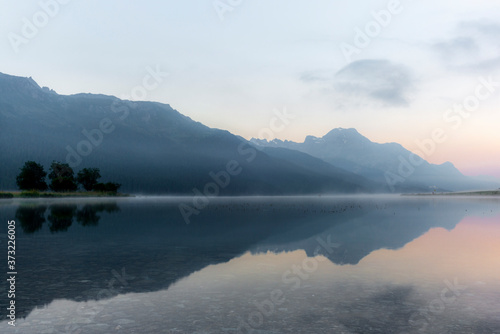 The mountains reflecting in the lake of Silvaplana in the Engadin valley at sunrise with the fog over the water