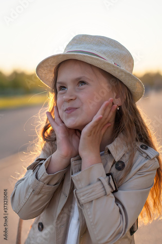 A little girl in a beige raincoat stands in the setting sun. Family walk in the Park