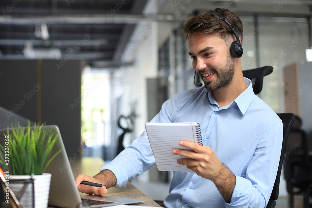 Smiling male call-center operator with headphones sitting at modern office, consulting online information in a laptop, looking up information in a file in order to be of assistance to the client. Smiling male call-center operator with headphones sitting at modern office, consulting online information in a laptop, looking up information in a file in order to be of assistance to the client.