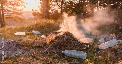 Time-lapse of a bunch of burning garbage left in the middle of a forest, during sunset, shot with a camera that's slowly moving to the right.