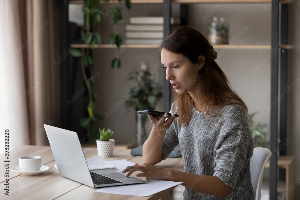 Focused young Caucasian woman sit at desk at home work on laptop record audio message on cellphone, millennial female use computer browsing, activate virtual digital voice assistant on smartphone Focused young Caucasian woman sit at desk at home work on laptop record audio message on cellphone, millennial female use computer browsing, activate virtual digital voice assistant on smartphone