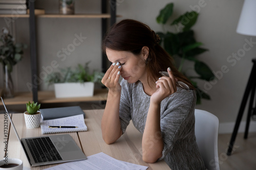 Exhausted young Caucasian woman work on laptop at home rub eyes suffer from blurry vision or dizziness, tired millennial female struggle with migraine or headache overwhelmed with computer job