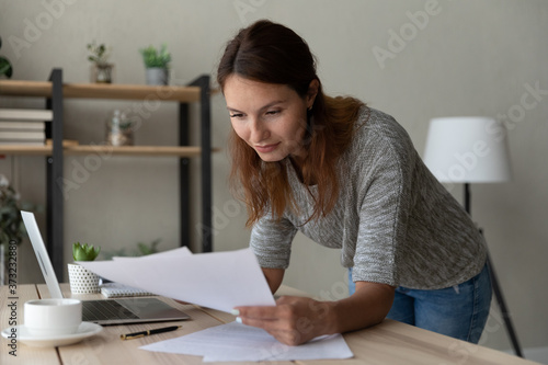 Focused young Caucasian businesswoman lean at table at home consider paper documents work on computer, millennial woman busy using laptop analyzing paperwork correspondence at workplace