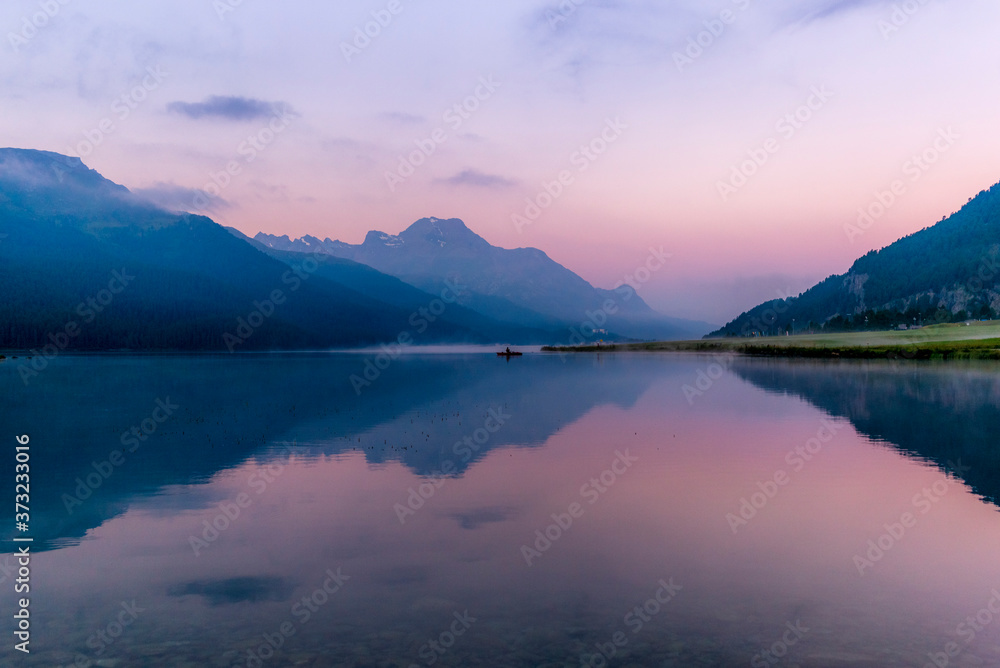 A fisherman rowing among the fog on the lake of Silvaplana in the Engadin valley at sunrise with mountains reflecting in the water
