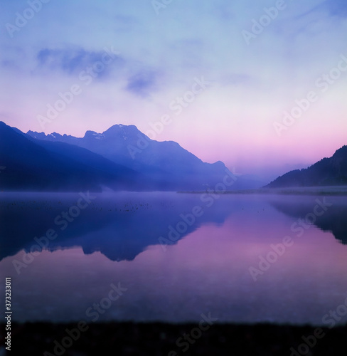 A fisherman rowing among the fog on the lake of Silvaplana in the Engadin valley at sunrise with mountains reflecting in the water © gdefilip