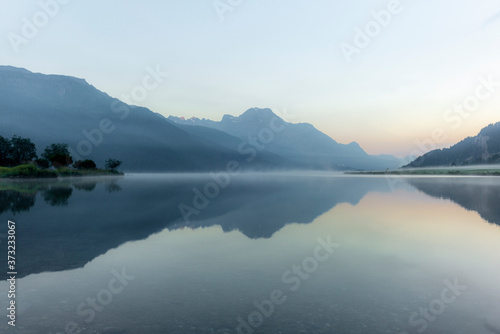The mountains reflecting in the lake of Silvaplana in the Engadin valley at sunrise with the fog over the water