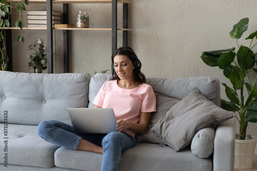 Young Arabic indian woman in headset sit on couch at home consult client or customer on webcam conversation on laptop. Millennial ethnic female in earphones talk on video call online. Young Arabic indian woman in headset sit on couch at home consult client or customer on webcam conversation on laptop. Millennial ethnic female in earphones talk on video call online.
