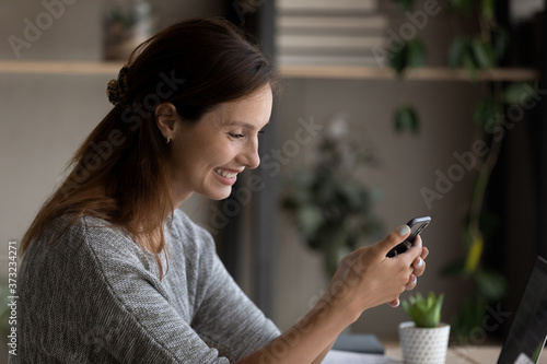 Smiling young Caucasian woman look at smartphone screen have fun texting messaging on gadget online, happy millennial female watch video or webinar on cellphone, use wireless internet connection