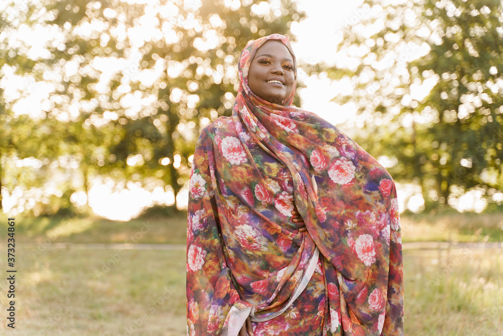 Muslim woman African ethnicity weared traditional colorful hijab smiles on the green meadow. Arabic islam religion clothes. Pretty black girl smiles