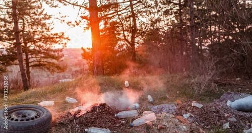 Time-lapse of a pile of burning garbage left in the middle of a forest, during sunset, shot with a camera that's slowly moving to the left.