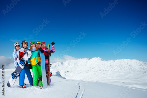 Group of young adults snowboarders stand on top of the mountain together over snow covered peaks on sunny weather on back