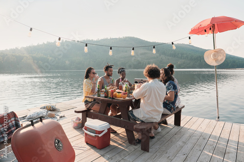 Canvas Print Group of friends sitting at the table and have dinner on a pier with beautiful l