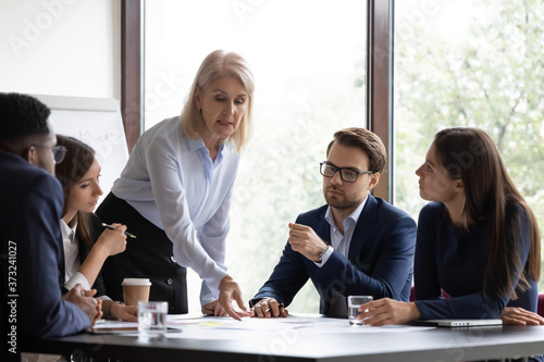 Aged woman experienced employee drawing attention of young colleagues to paper documents on office meeting, mature female trainer or leader pointing diverse staff to important detail in business plan