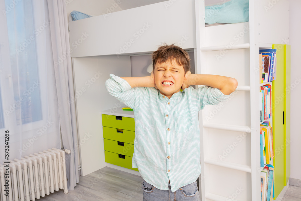 Little autistic boy with strong negative face expression closing ears and grins teeth Little autistic boy with strong negative face expression closing ears and grins teeth