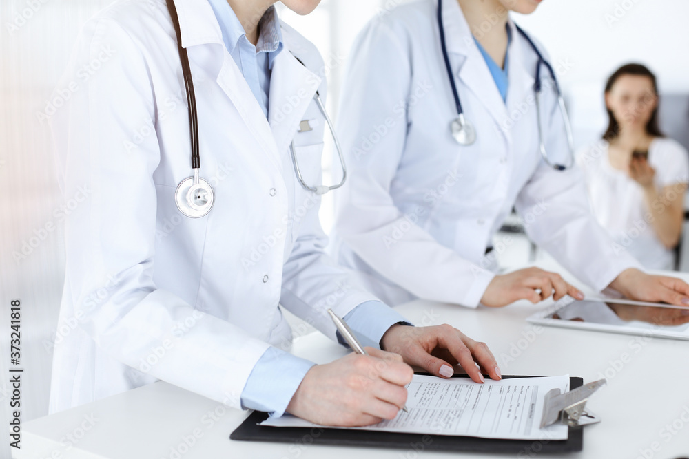 Unknown woman-doctors at work with patient at the background. Female physicians filling up medical documents or prescription while standing in sunny hospital reception desk, close-up. Health care Unknown woman-doctors at work with patient at the background. Female physicians filling up medical documents or prescription while standing in sunny hospital reception desk, close-up. Health care