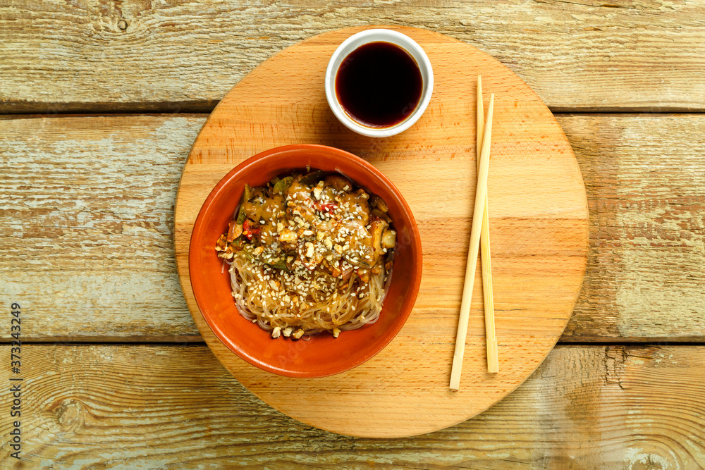 Funchoza with vegetables and mushrooms in a clay plate on a round stand next to chopsticks and soy sauce. Funchoza with vegetables and mushrooms in a clay plate on a round stand next to chopsticks and soy sauce.