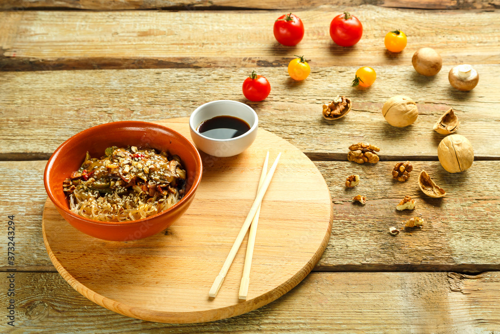 Funchoza with vegetables and mushrooms in a clay plate on a round stand next to chopsticks and soy sauce next to vegetables Funchoza with vegetables and mushrooms in a clay plate on a round stand next to chopsticks and soy sauce next to vegetables