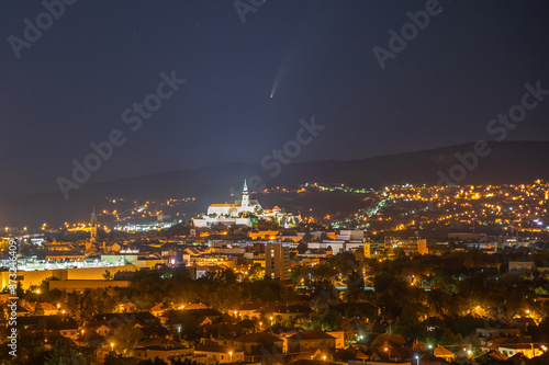 Comet Neowise in the night sky over the illuminated city and mountains in the background