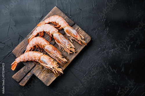 Seafood shrimps on wooden cutting board on black concrete surface, flat lay with copy space