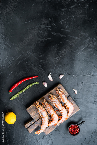 Seafood shrimps on wooden cutting board with spices, pepper and lemon on black concrete surface, flat lay with copy space
