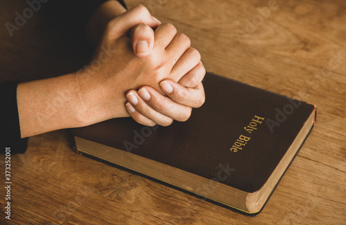 Woman with Bible praying, hands clasped together on her Bible on wooden table.