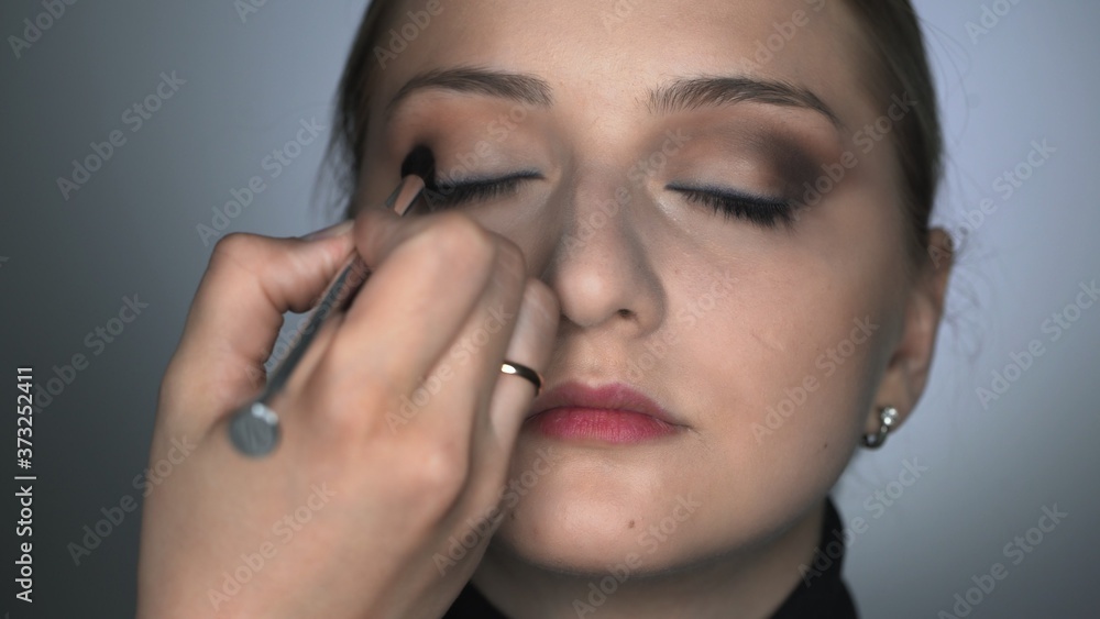 Front view of makeup artist making professional make-up for young woman in beauty studio. Make up Artist uses brush to applies shadow on eyelid
