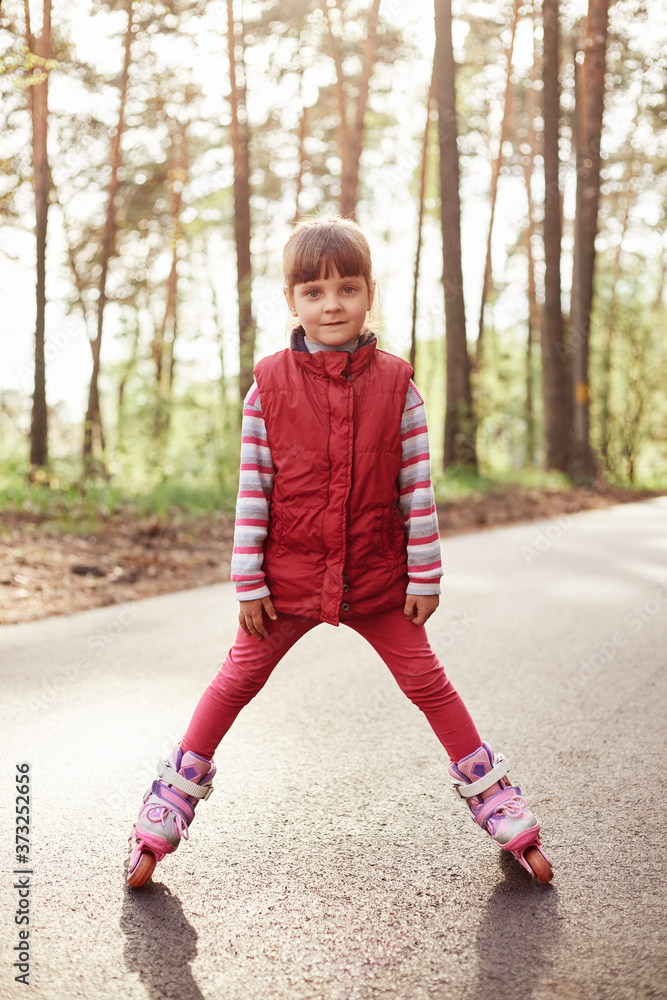 Fototapeta premium Cute girl posing on road in forest, wearing casual shirt and rolling skate, looking directly at camera, spending time in open air, being photographed by mother.