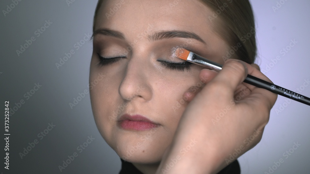 Makeup artist making professional make-up for young woman in beauty studio. Make up Artist uses brush to applies glitter on eyes