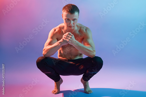 Canvas Print Handsome sportive man meditating in yoga asanas, squatting and standing on legs, looks concentrated, posing topless isolated over neon background