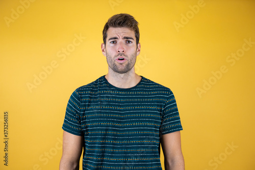 Handsome young man standing over insolated yellow background looking at the camera surprised