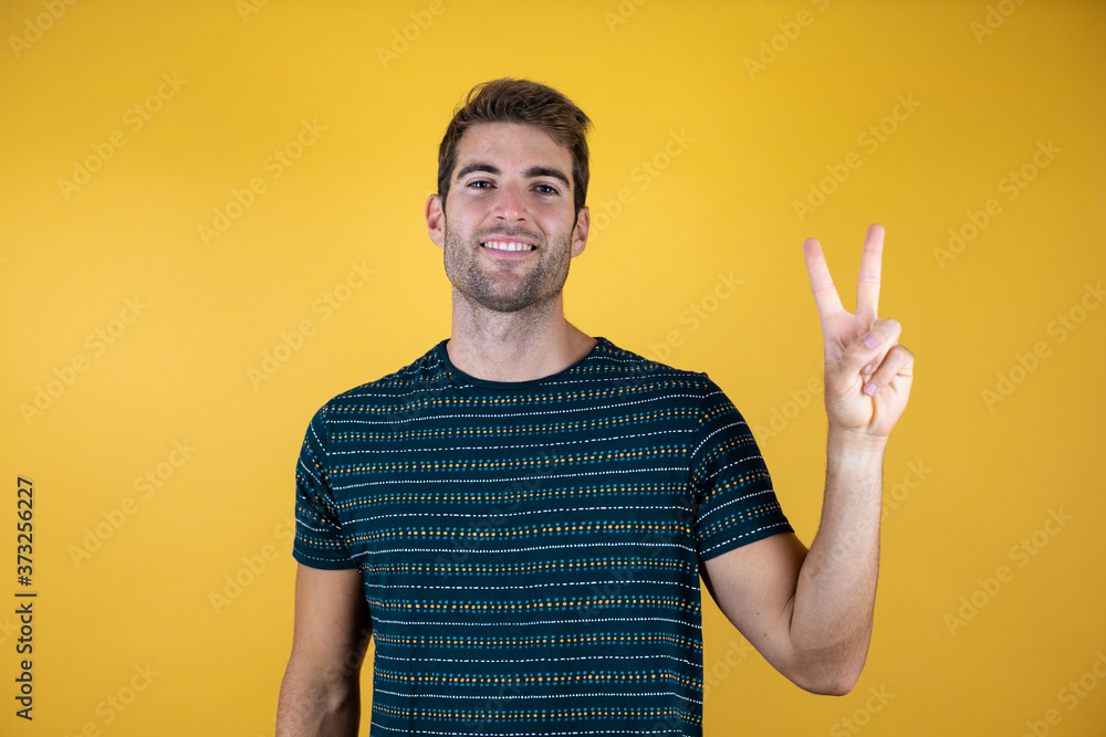 Young handsome man wearing casual blue t-shirt over yellow isolated background smiling with happy face winking at the camera doing victory sign. Number two. Young handsome man wearing casual blue t-shirt over yellow isolated background smiling with happy face winking at the camera doing victory sign. Number two.