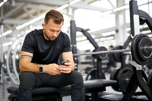young tired fitness man using smartphone in gym. male in black sportswear have rest after exercises, look at scren of smartphone, chat with someone