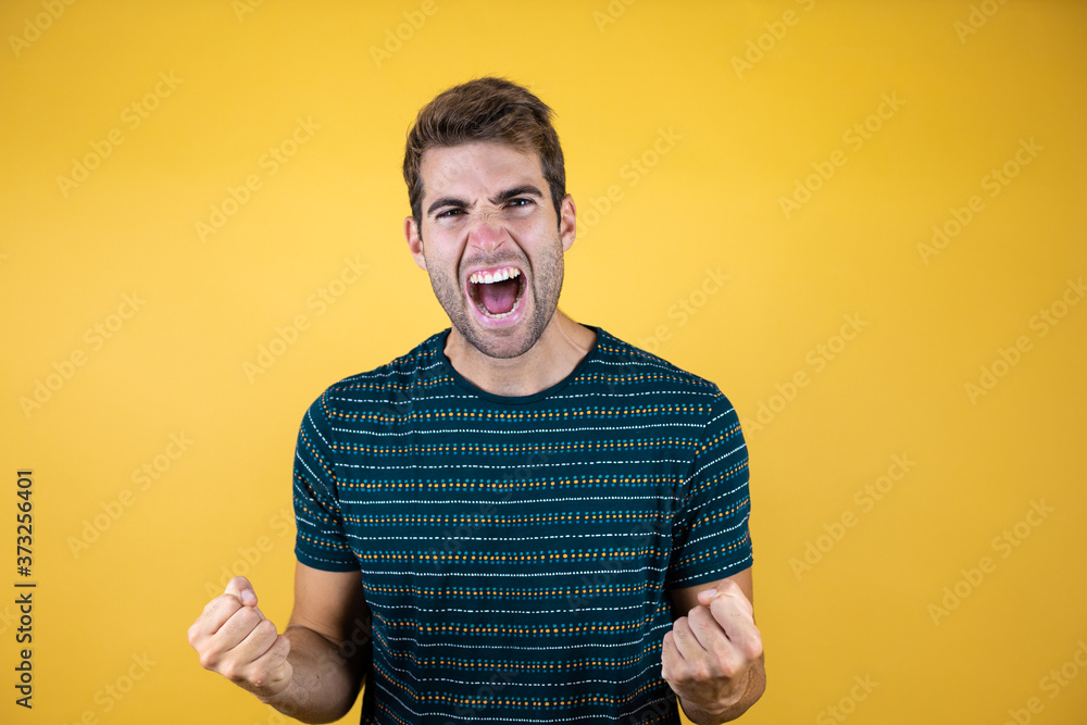 Young handsome man wearing blue t-shirt over yellow isolated background very happy and excited doing winner gesture with arms raised, smiling and screaming for success. Celebration concept. Young handsome man wearing blue t-shirt over yellow isolated background very happy and excited doing winner gesture with arms raised, smiling and screaming for success. Celebration concept.