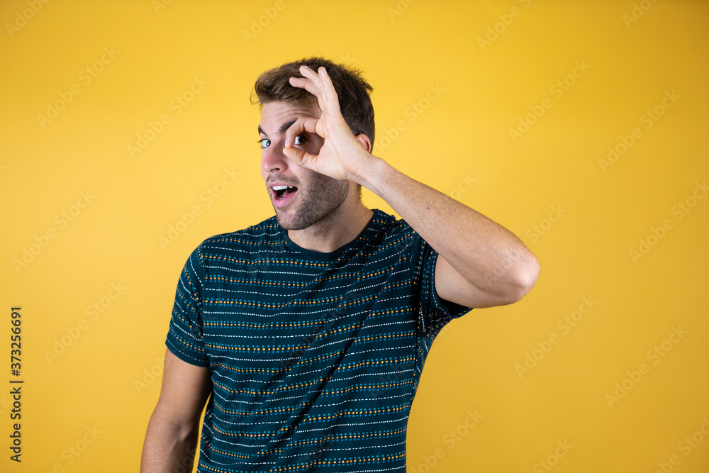 Handsome man standing over isolated yellow background doing okay gesture shocked with surprised face, eye looking through fingers Handsome man standing over isolated yellow background doing okay gesture shocked with surprised face, eye looking through fingers